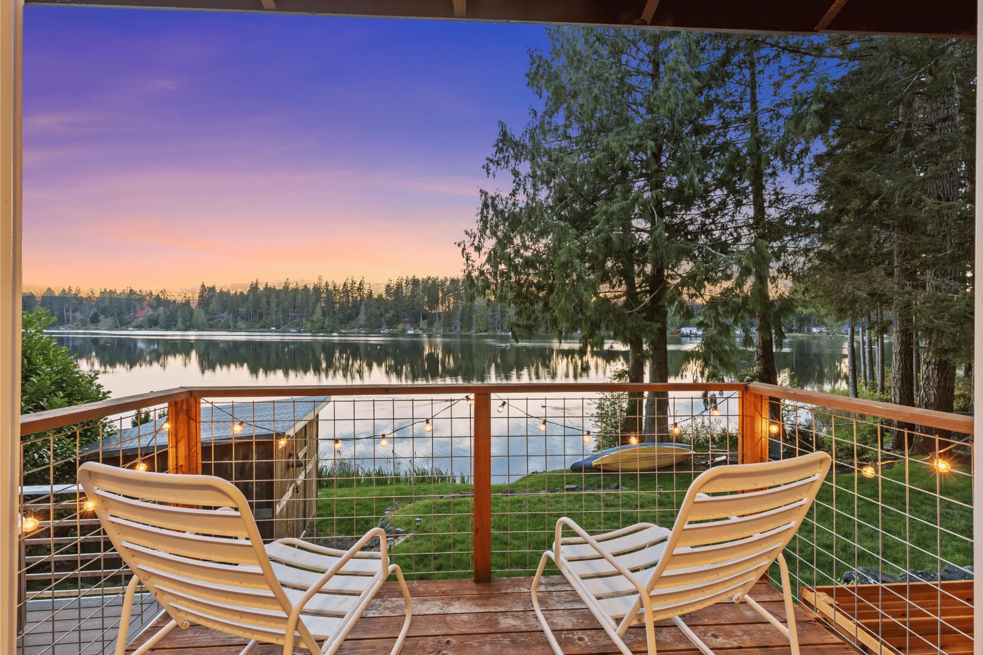 Two lounge chairs on the deck of Cutty Cabin overlooking Trails End Lake at twilight, string lights glowing near Hood Canal